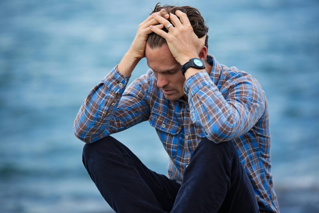 alerte scam arnaque A man in a plaid shirt sits by the water looking distressed, symbolizing stress.